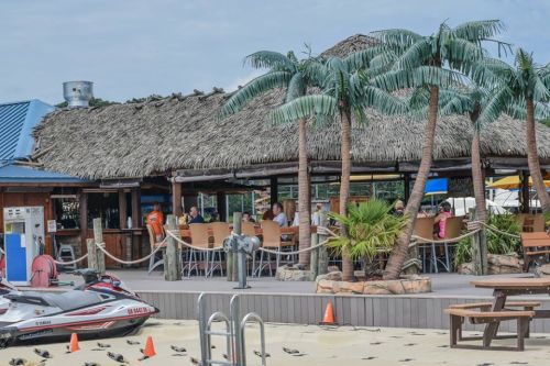 A tropical beachside restaurant with palm trees, outdoor seating, and jet skis nearby.