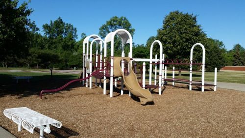 A playground with a brown slide, climbing structures, and a sandy area, surrounded by trees and clear blue skies.