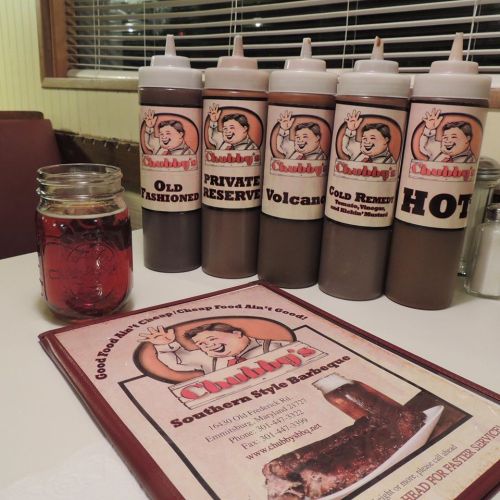 A table with a menu and five bottles of barbecue sauce, alongside a jar of drink, in a cozy restaurant setting.
