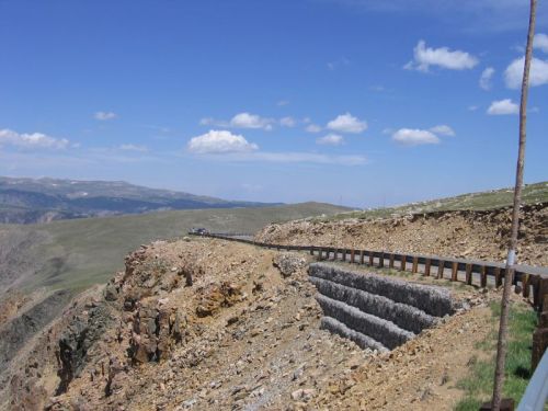 A winding road along a rocky cliff with a clear blue sky and distant mountains in the background.