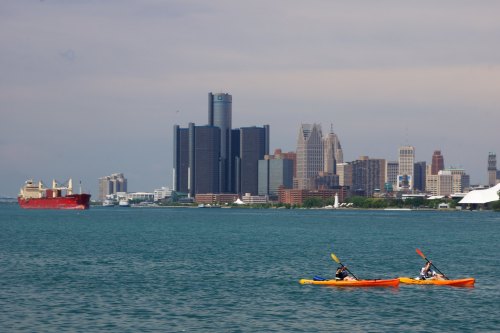 Two kayakers paddle in the foreground, with a city skyline and a cargo ship in the background on a calm waterway.