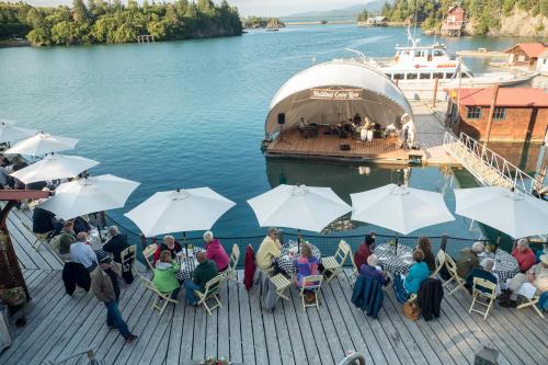 A waterfront dining area with umbrellas, people enjoying a meal, and a stage for live music by the water.