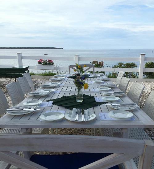 A beautifully set outdoor dining table by the water, with flowers and plates, under a cloudy sky.