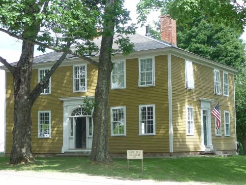 Historic yellow house with white trim, large windows, and an American flag, surrounded by trees. Samuel Reed Hall House sign.