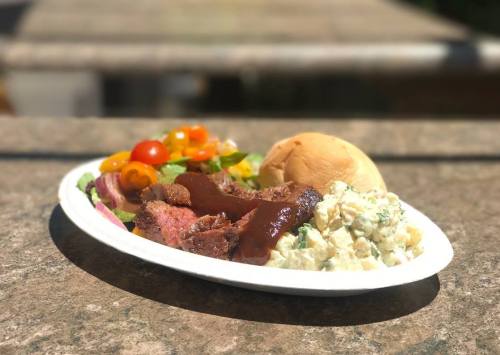A plate of grilled meat, potato salad, mixed salad with cherry tomatoes, and a soft roll, set outdoors.