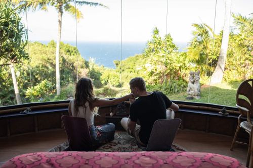 A couple sits together in a cozy room, gazing at a scenic ocean view through large glass windows.
