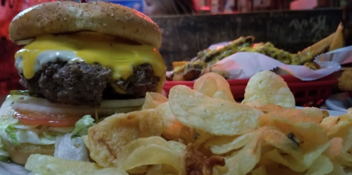 A close-up of a cheeseburger with lettuce, tomato, and chips, with a side of fries in the background.