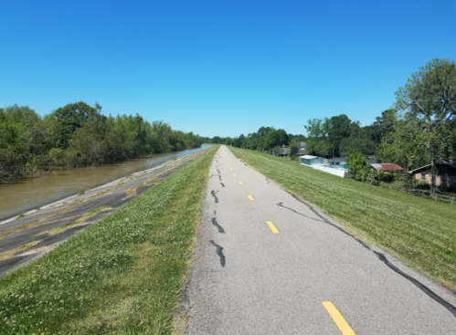 A paved path runs alongside a canal, with green grass and trees on one side and houses on the other under a clear blue sky.