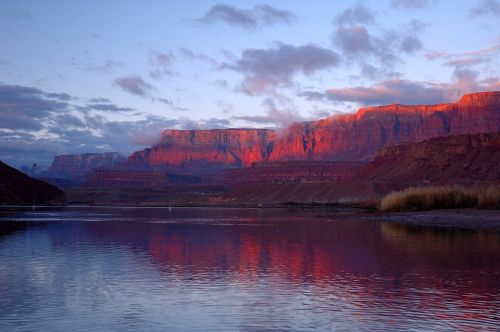 Sunset over the Grand Canyon, with vibrant red cliffs reflecting in the calm river below and clouds in the sky.