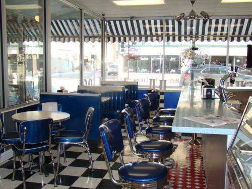 A retro diner interior with blue booths, chrome stools, and a checkered floor, featuring large windows and striped awnings.