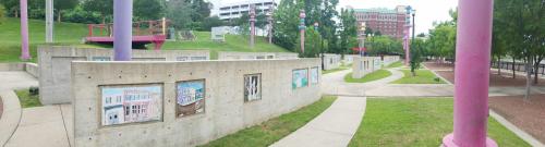 A panoramic view of a park featuring colorful pillars and art displays on concrete walls amidst green grass and trees.