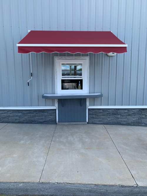 A small service window with a red awning on a gray metal building, featuring a stone base and concrete walkway.