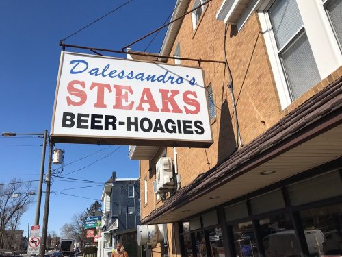 Sign for Dalessandro's featuring "Steaks," "Beer," and "Hoagies" against a brick building backdrop.