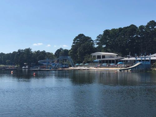 A serene lake view featuring a beach area, water slides, and a modern building surrounded by trees.
