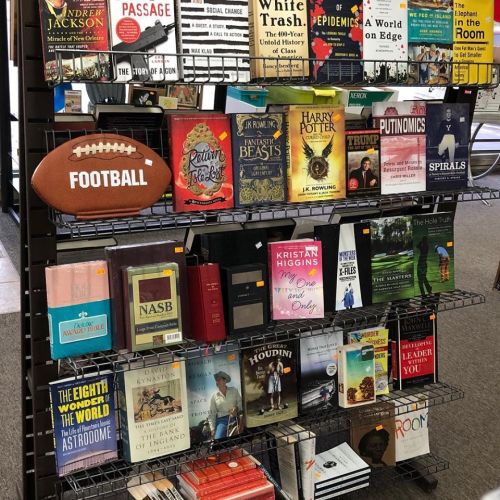 A display of books on a rack, featuring titles by various authors, with a football placed on top.