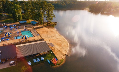 Aerial view of a lakeside area with a pool, sandy shore, and kayaks, surrounded by trees and reflecting water.