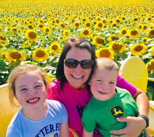 A woman and two children smile together in a vibrant sunflower field, surrounded by bright yellow flowers.
