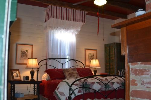 Cozy bedroom with a quilted bed, two lamps, and a window draped with sheer curtains. Warm, inviting decor.