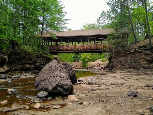 A wooden covered bridge spans a rocky creek surrounded by lush green trees and a serene landscape.