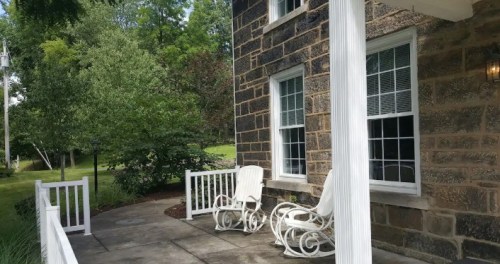 A porch with two white rocking chairs, surrounded by greenery and a stone wall, creating a peaceful outdoor space.