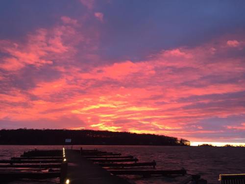 A vibrant sunset paints the sky in shades of pink and orange over a calm lake, with a wooden pier extending into the water.