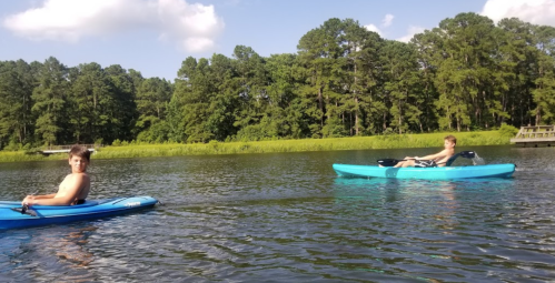 Two boys kayaking on a calm lake surrounded by green trees under a partly cloudy sky.