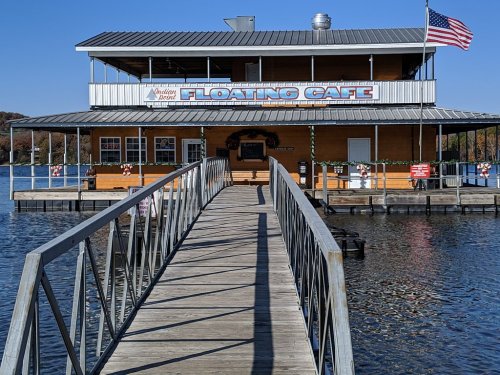 A wooden dock leads to a two-story floating café with festive decorations and an American flag.