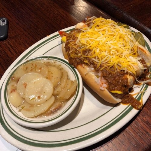 A loaded hot dog topped with chili and cheese, served with a side of sliced potatoes in a creamy sauce.