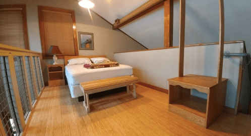 Cozy loft bedroom with a bed, wooden bench, and lamp, featuring warm wood accents and natural light.