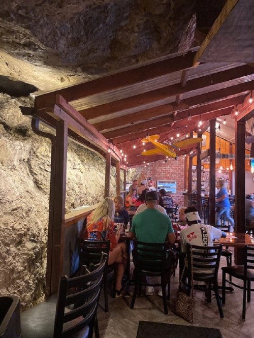 Cozy restaurant interior with stone walls, wooden beams, and patrons dining at tables under warm lighting.