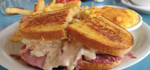A close-up of a grilled sandwich filled with meat and sauce, served with fries and mac and cheese on the side.