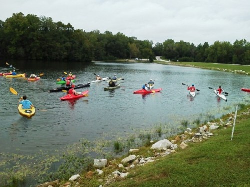 A group of kayakers paddles on a calm lake surrounded by greenery and a cloudy sky.