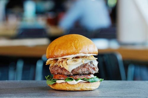 A juicy burger with lettuce, tomato, onions, and crispy toppings, served on a soft bun against a blurred background.