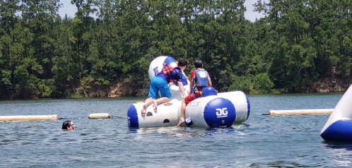 Children play on inflatable water structures in a lake, with trees in the background and one person swimming nearby.