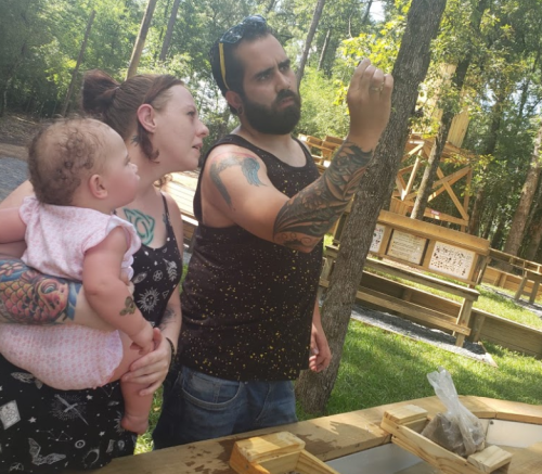 A woman holds a baby while a man examines something in a wooded outdoor setting. All are engaged and focused.