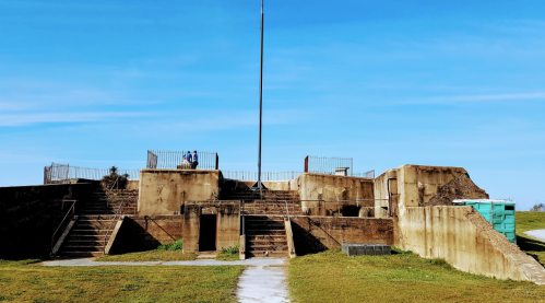 Historic concrete structure with stairs, a flagpole, and two people sitting on top, set against a clear blue sky.