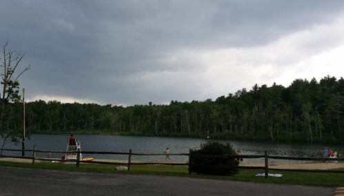A cloudy day at a lake with people walking along the shore and trees lining the background.