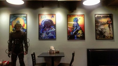 A dimly lit restaurant featuring four colorful sci-fi posters on the wall and a life-sized robot figure standing nearby.