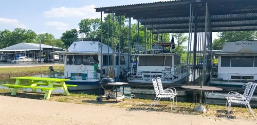 Two houseboats docked under a shelter, with picnic tables and chairs nearby, surrounded by greenery and blue skies.