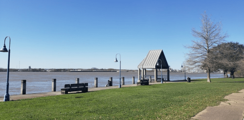 A riverside park with benches, a gazebo, and a clear blue sky. The water is calm and the grass is green.