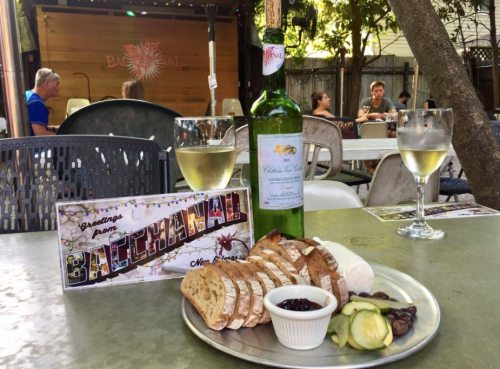 A plate of bread, cheese, and pickles with a bottle of beer, set on a table at Bacchanal, with people in the background.