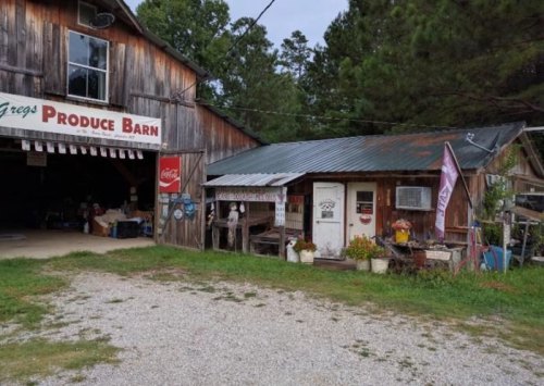 A rustic produce barn with a wooden exterior, metal roof, and a small attached shop, surrounded by greenery.