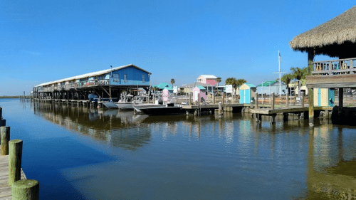 Colorful beach huts line a calm waterway with boats docked, under a clear blue sky.