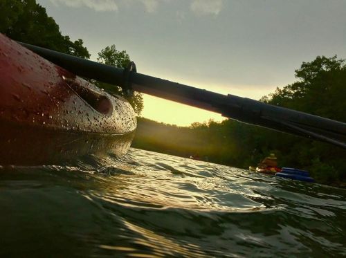 A close-up of a kayak on water at sunset, with raindrops on the surface and trees in the background.