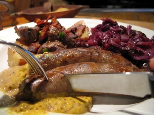 A plate of sausages with mustard, red cabbage, roasted vegetables, and a fork and knife ready to eat.