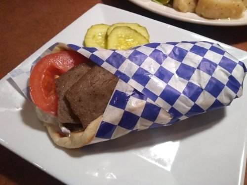 A wrapped sandwich with meat, tomato, and sauce, served with pickles on a plate.
