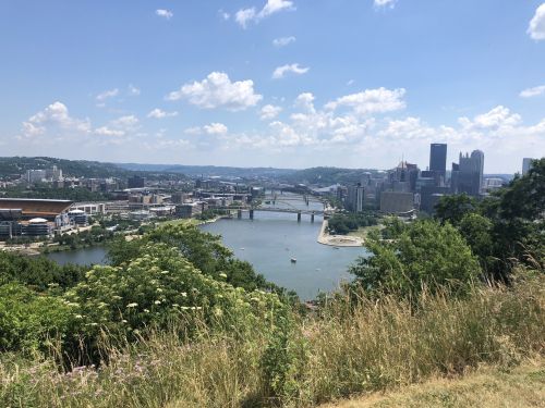 A panoramic view of Pittsburgh's skyline, rivers, and green hills under a blue sky with fluffy clouds.