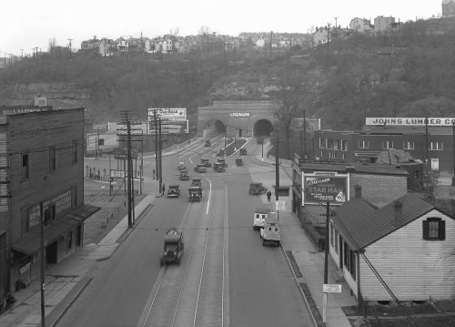 Black and white image of a road with cars, tunnels, and buildings, set in a hilly urban landscape.