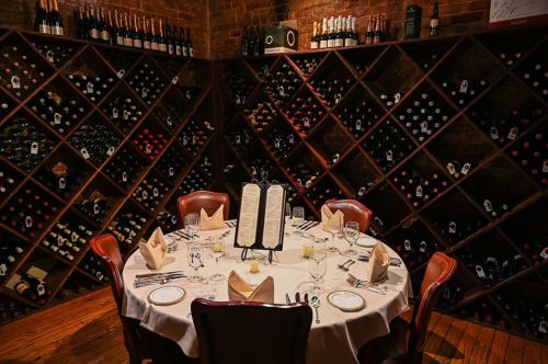 A round dining table set for a meal, surrounded by a wall of wine bottles in a cozy restaurant setting.