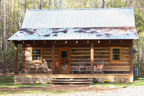 A rustic log cabin with a metal roof, featuring a front porch with rocking chairs and surrounded by trees.